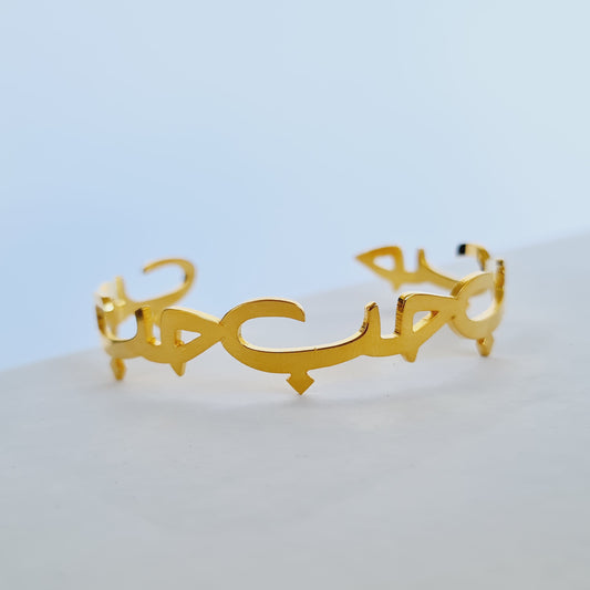 A gold-colored cuff bracelet with an adjustable fit and intricate designs, displayed against a light background.سوار حب مفتوح (كَفّ)
